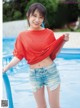 A woman in a red shirt and denim shorts posing by a pool.